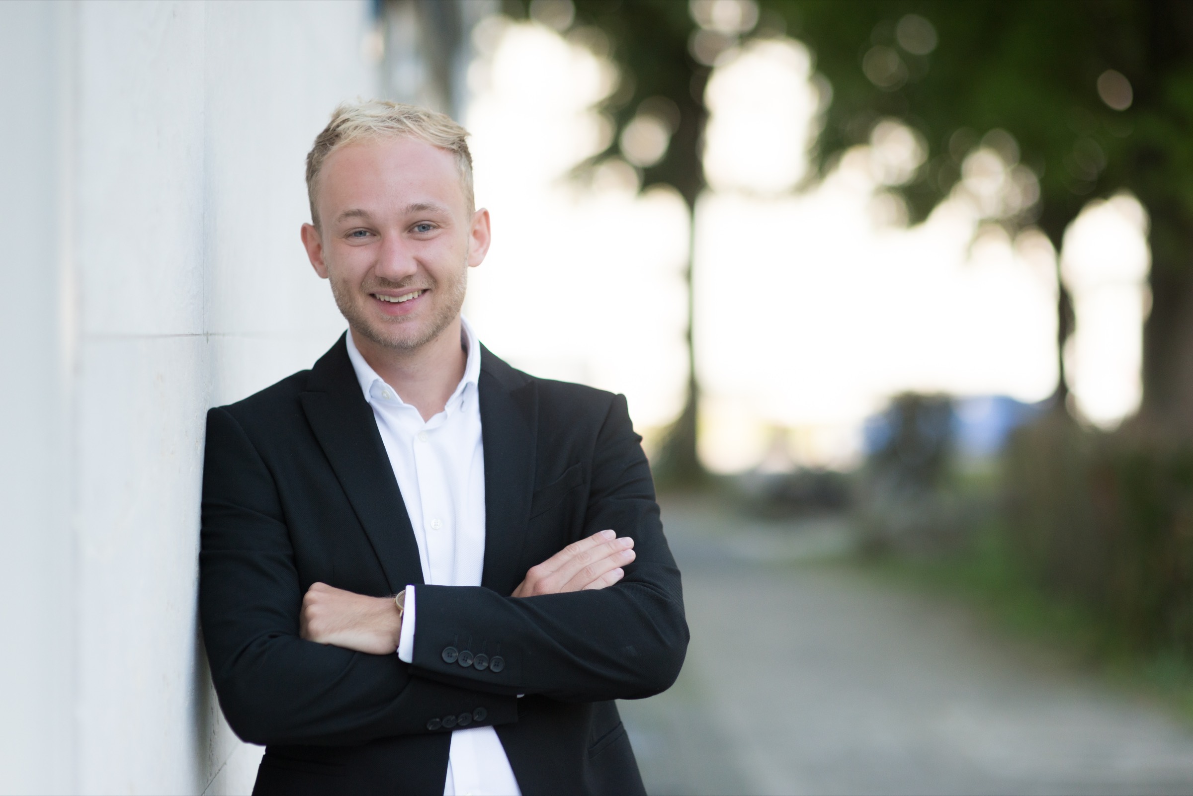 Johannes Lang, lächelnd, verschränkte Arme, im Hintergrund eine unscharfe Straße mit Bäumen